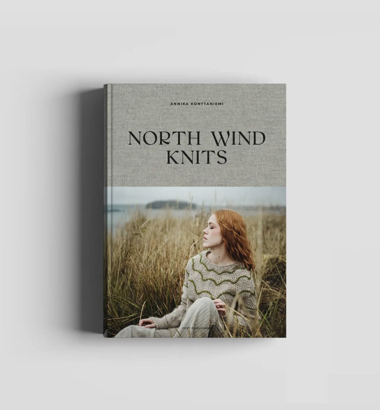 Boekomslag van "North Wind Knits" door Annika Konttaniemi. Op de omslag zit een vrouw met rood haar in hoog kustgras, gekleed in een lichtgrijze handgebreide trui met groen patroon. Op de achtergrond zijn water en een eiland zichtbaar.