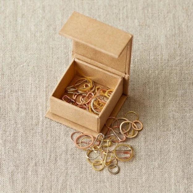 Vierkant kartonnen doosje met magneetsluiting van CocoKnits gevuld met ronde en opening stitch markers in koper-, goud- en zilverkleur