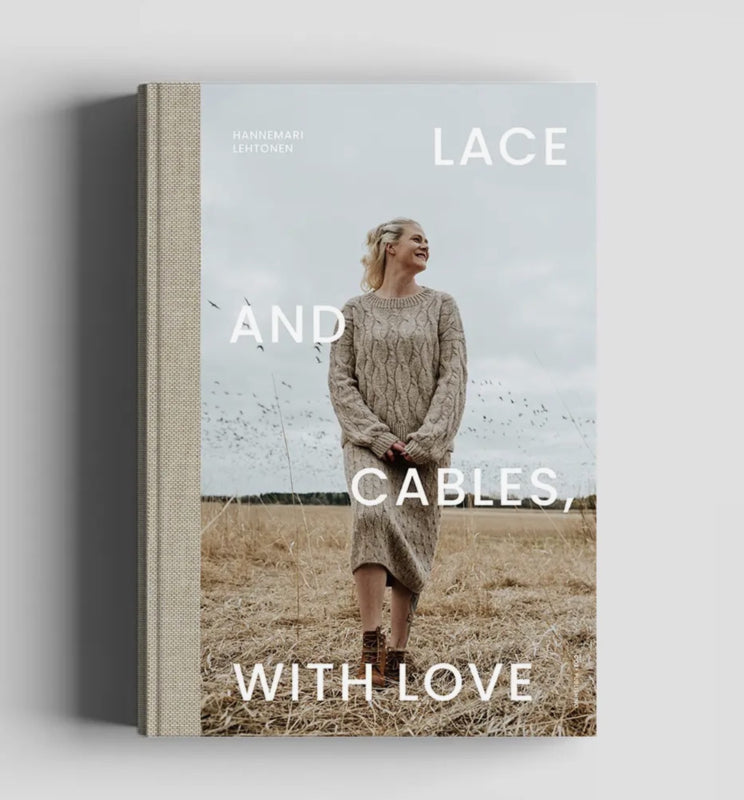 Boekomslag van "Lace and Cables with Love" door HanneMari Lehtonen. Op de omslag staat een vrouw in een lichtgekleurde, handgebreide kabeltrui-jurk in een droog grasveld onder een bewolkte lucht. Het boek heeft een linnen rug en een lichtblauwe omslag.